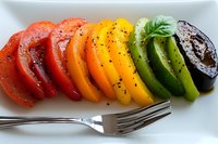 Fresh colorful heirloom tomato slices arranged in rainbow gradient from red to purple, seasoned with black pepper and herbs, served on white plate with fork.