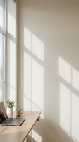 Minimalist home office workspace with natural sunlight casting window shadows on neutral beige wall. Wooden desk with laptop and plant creates calm productive environment.