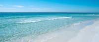 Crystal clear turquoise ocean waves gently washing onto pristine white sand beach under bright blue sky with fluffy white clouds on sunny tropical day.