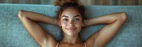 Young mixed race woman with curly hair relaxing on turquoise couch, hands behind head, expressing joy and contentment in casual home setting.