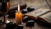 Mystical candle flame illuminates ancient book with ornate chalice and smooth stones on rustic wooden surface creating atmospheric spiritual scene.
