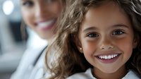 Happy young girl with curly hair smiling at camera while mother stands behind her in bright indoor setting. Family bonding moment captured in natural light.