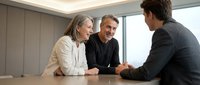 Mature woman with gray hair and two men in business casual attire discussing strategy at wooden table in modern office with natural light from windows.