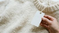 Blank white tag attached to beige knit sweater held in hand, showcasing clothing label for branding, fashion retail, and garment care instructions.