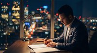 Professional businessman working late at night in modern office with city lights background, focused on paperwork and documents at wooden desk with lamp.