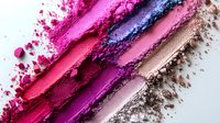 Crushed makeup eyeshadow and powder swatches in pink, purple, and brown shades creating diagonal lines with scattered powder texture on white background.