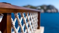 Nautical rope railing detail on wooden deck with blurred ocean and rocky coastline background, featuring twisted white marine cordage in decorative pattern.