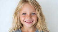 Young Caucasian girl with blonde wavy hair and bright blue eyes smiling at camera against white background, conveying happiness and innocence.