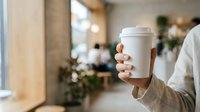 Woman holding white takeaway coffee cup in modern cafe interior with blurred background, suitable for beverage, lifestyle and hospitality marketing materials.