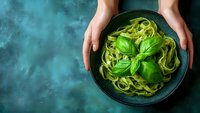 Hands holding bowl of fresh spinach pasta with basil leaves on teal background, perfect for food blogs and recipe websites.