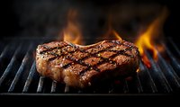 Juicy beef steak grilling on flaming barbecue grill with perfect sear marks and fire flames in dark moody atmosphere, close up view of premium meat cooking.