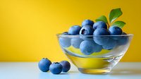 Fresh blueberries in glass bowl with green leaves against bright yellow background, perfect for food blogs and healthy eating content.