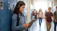Young Caucasian female student using digital tablet in school hallway near blue lockers with classmates walking in background during break time.