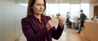 Middle-aged Caucasian businesswoman in burgundy blazer examining wooden puzzle piece in modern office environment for problem-solving concepts.