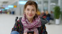 Young Caucasian woman wearing floral winter jacket and pink knitted scarf smiling at airport terminal, expressing joy and positive emotions during travel.