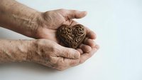 Elderly hands gently holding ornate wooden heart symbol representing love, care, protection and compassion in cupped palms on white background.