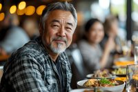 Mature Asian man with gray beard smiling while dining at restaurant, wearing casual plaid shirt. Warm bokeh lighting creates welcoming ambiance for lifestyle and hospitality content.