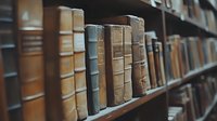 Vintage leather-bound books arranged on wooden library shelf, showing aged spines in various neutral tones and textures, creating atmospheric academic ambiance.