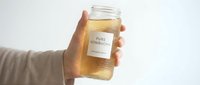 Hand holding glass jar of golden kombucha with white label against clean background. Healthy fermented probiotic drink for wellness and nutrition.