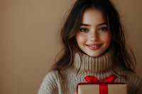 Young woman with dark hair and bright smile holding gift box with red bow in beige turtleneck sweater against brown background. Cozy holiday portrait.