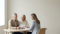 Mature man and woman with gray hair reviewing documents with younger woman at wooden table during business meeting or consultation.