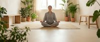 Young woman practicing meditation in peaceful home studio surrounded by lush green plants and natural light creating serene wellness atmosphere.