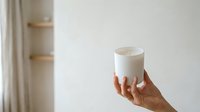 Woman's hand holding lit white scented candle against neutral interior background. Suitable for wellness, home decor, relaxation, and lifestyle marketing materials.