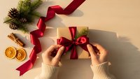 Hands wrapping Christmas gift with red ribbon on beige background with festive decorations, pine branches, cinnamon sticks and dried orange slices.