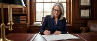 Mature woman with gray hair and glasses wearing dark blazer writing at wooden desk in traditional law office with bookshelves and window.