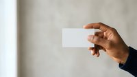 Cropped hand of a man with dark skin tone holding a blank white business card against a neutral gray wall background, ready for branding and design mockups.