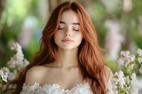 Young Caucasian woman with long red hair and closed eyes in white lace dress surrounded by spring flowers for wellness and beauty concepts.