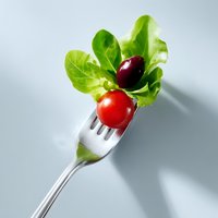 Fresh salad ingredients on silver fork with cherry tomato, black olive and green lettuce leaves against light background with dramatic shadows.