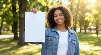 Young African American woman with curly hair holding white shopping bag in sunny park, smiling at camera wearing denim jacket for retail and lifestyle concepts.