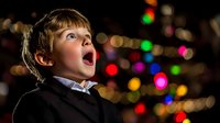 Young Caucasian boy in formal attire expressing amazement with open mouth against blurred colorful Christmas lights background creating magical holiday atmosphere.
