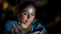 Young Latin American woman in traditional colorful dress and jewelry looking at mobile phone screen with gentle smile, illuminated by screen light in dark setting.