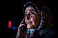 Middle Eastern senior woman wearing traditional hijab headscarf talking on mobile phone at night, illuminated by city lights with bokeh background.