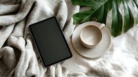 Digital tablet with coffee cup on cozy beige blanket next to tropical monstera leaf, creating relaxed home office atmosphere.