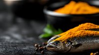 Fresh whole fish covered in golden turmeric powder with herbs on dark surface for culinary preparation and cooking concepts.