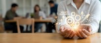 Businessman holding glowing gears and growth icons above wooden table with blurred team in background, representing business expansion and development strategy.
