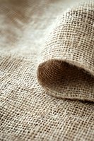 Natural burlap fabric with rolled edge showing woven texture and fibers in warm beige tones, suitable for rustic design backgrounds and eco-friendly packaging concepts.