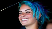 Young woman with vibrant blue hair laughing joyfully against dark background, showing genuine happiness and positive emotion in dramatic lighting.