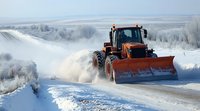 Orange tractor with snow plow clearing snowy rural road during winter storm, creating dramatic snow spray against frosty white landscape and cloudy sky.