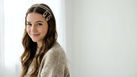 Young brunette woman with wavy hair wearing decorative hair clips and cozy beige sweater smiling at camera in bright natural light interior setting.