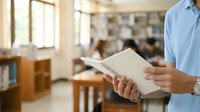 Man in light blue shirt holding open book in library with blurred bookshelves and people in background, suitable for education, learning, and knowledge concepts.
