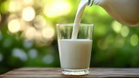 Fresh milk being poured into clear glass on wooden surface with natural green bokeh background for healthy breakfast and dairy nutrition concepts.