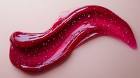 Glossy red cosmetic smear with sparkly glitter particles on pink background, macro shot showing texture and shine for beauty and makeup industry applications.