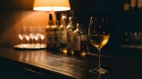 Glass of white wine on wooden bar counter with warm ambient lighting and blurred bottles in background creating cozy restaurant atmosphere.