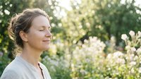 Middle-aged Caucasian woman with brown hair in updo, wearing gray shirt, smiling peacefully outdoors among blooming white flowers and green foliage. Suitable for wellness, lifestyle, and nature themes.