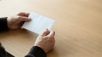 Hands of an adult male with medium skin tone holding blank white paper on wooden table surface, suitable for mockups, presentations, and business communications.
