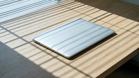 Laptop computer with silver metallic finish resting on wooden table surface with natural sunlight casting geometric shadows, suitable for technology, business, and workspace design projects.